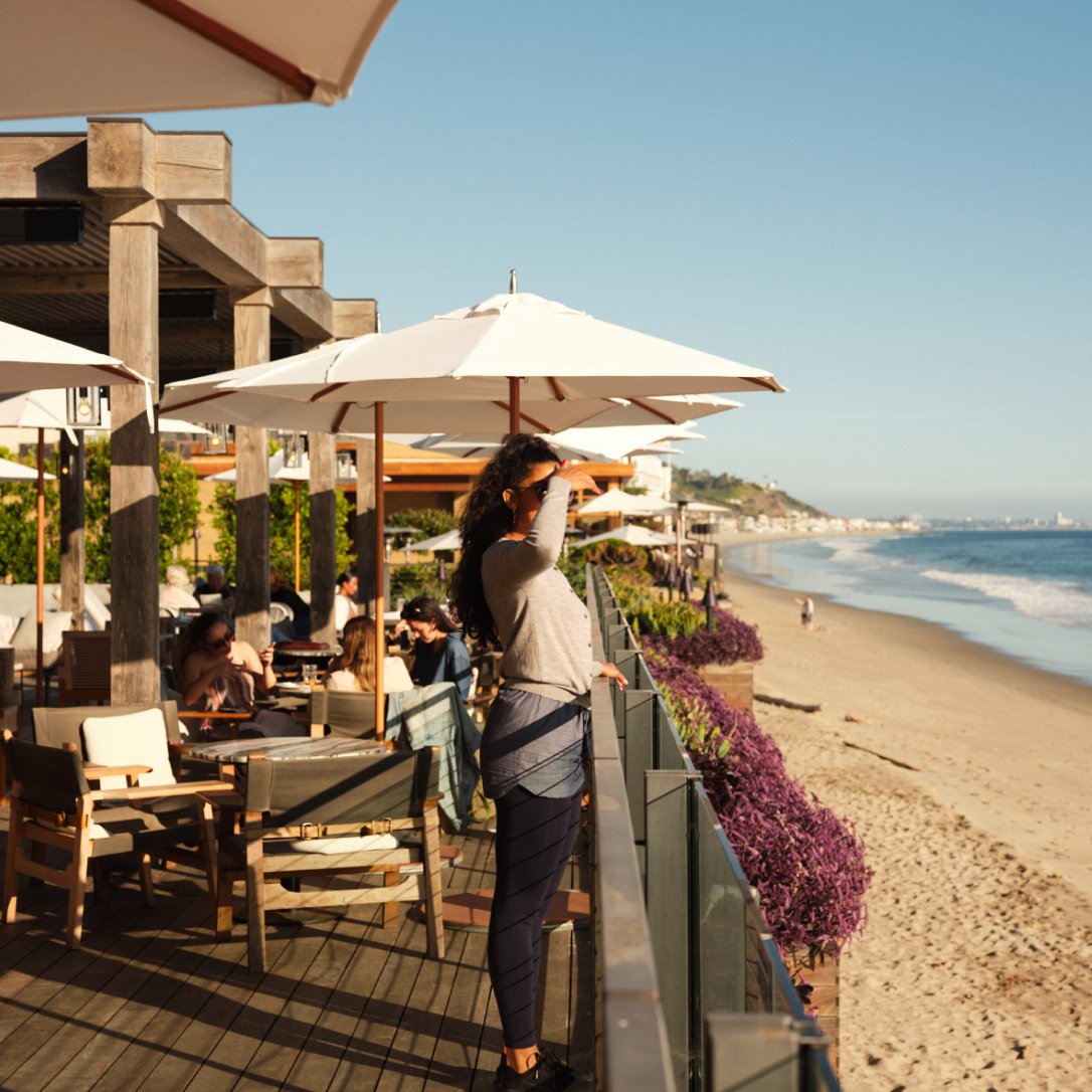 People on a terrace with dining tables overlooking a beach and the sea.