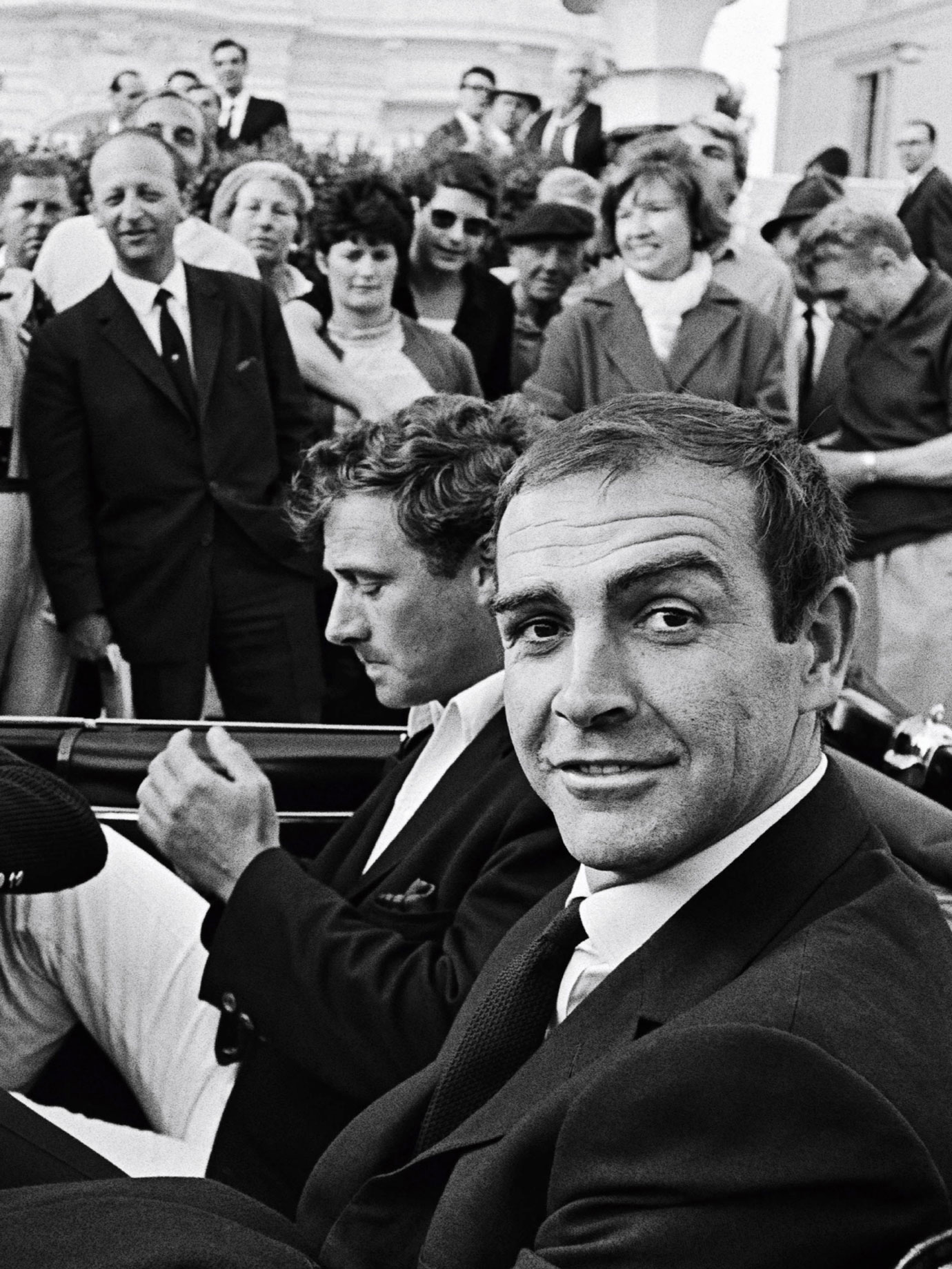 A man smiling at the camera in the back of an open top car as people look on in the 1960s.