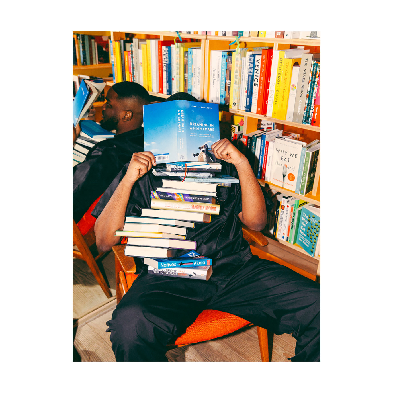 A man covered in a pile of books next to a yellow bookcase.
