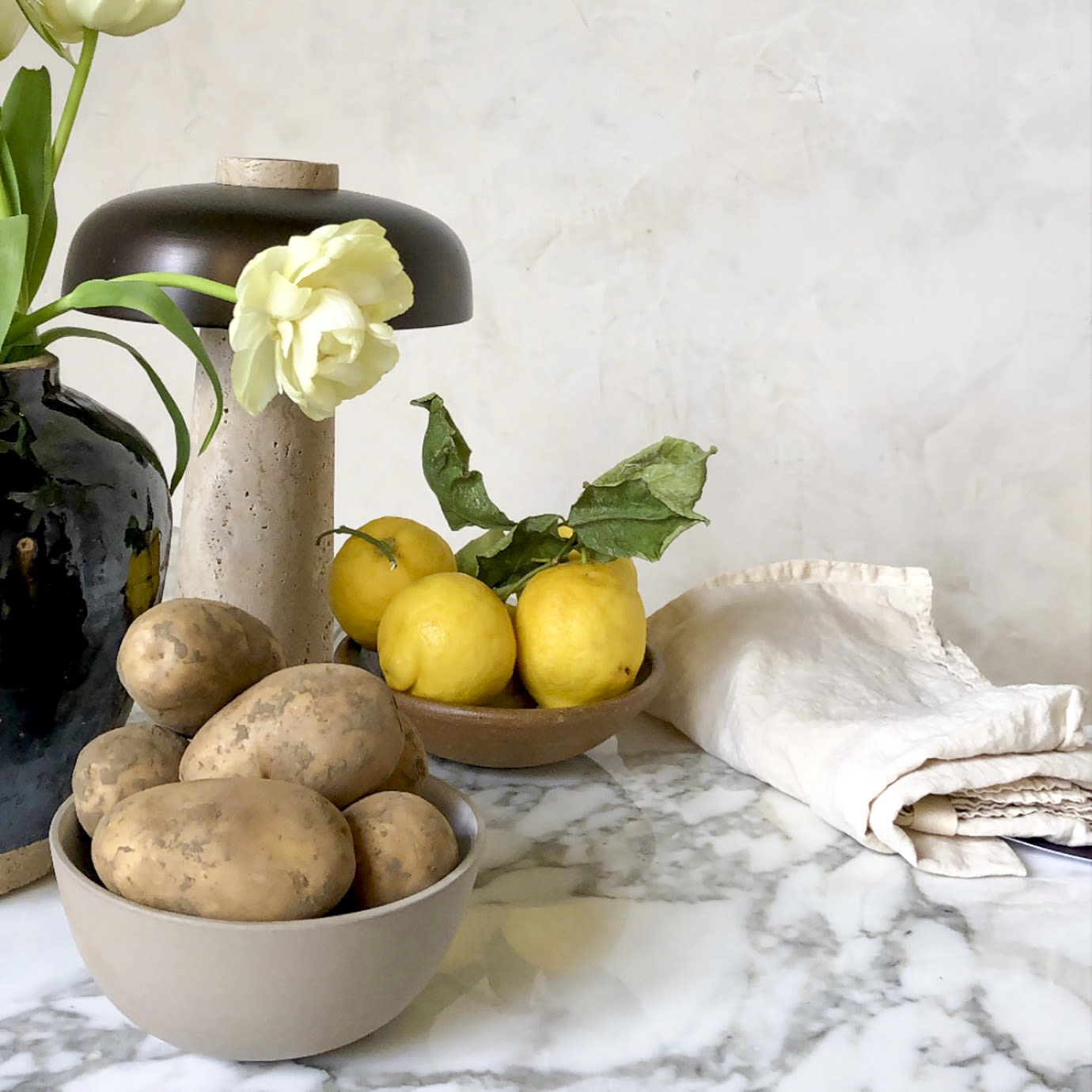 A bowl of lemons next to potatoes and flowers.