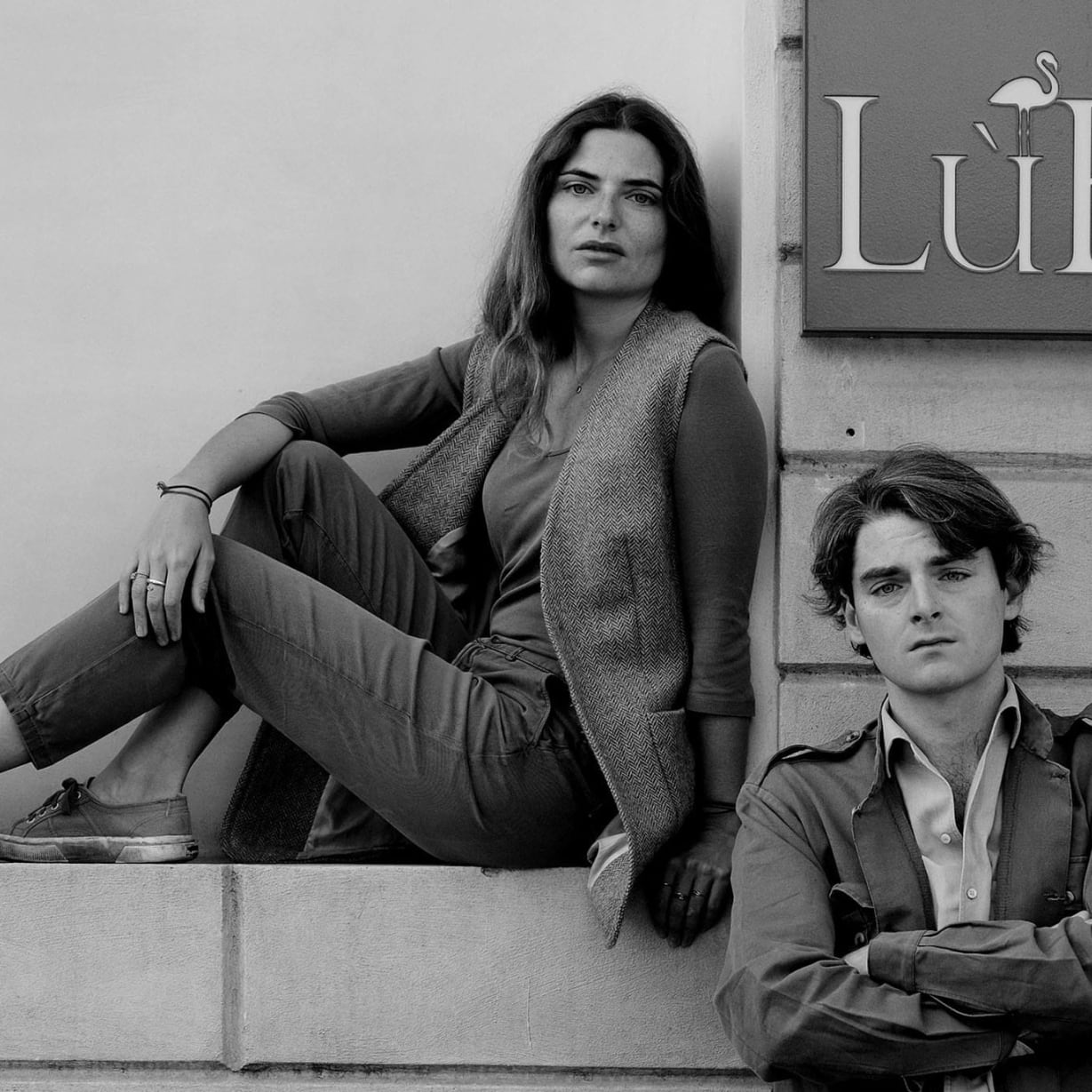 a woman sat in a building alcove and a man next to her with arms crossed
