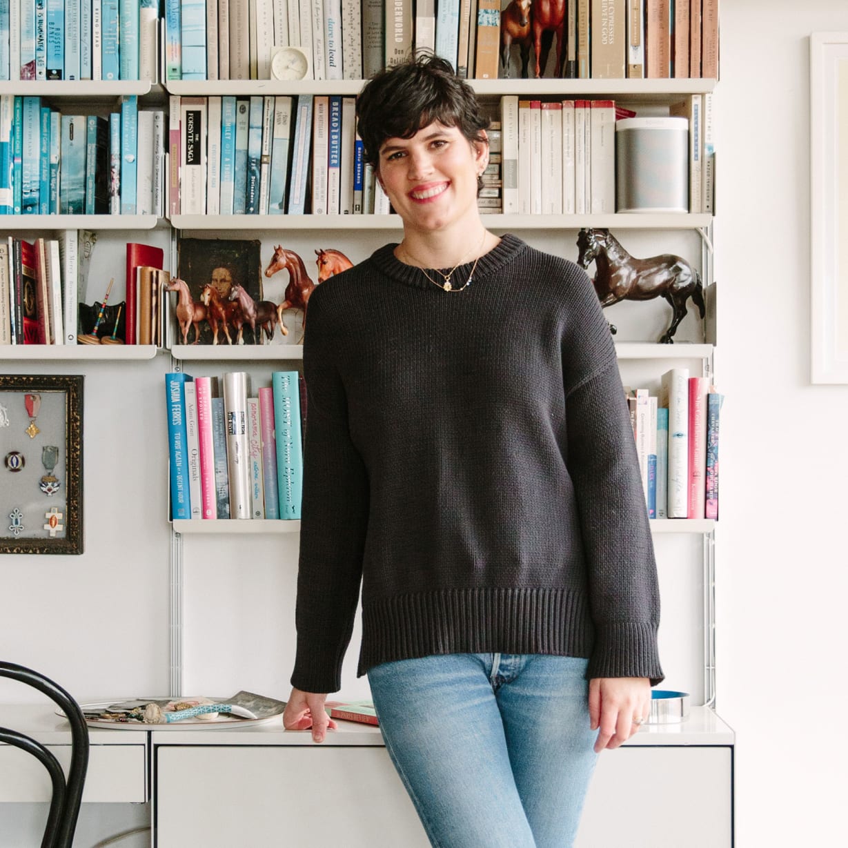 A woman standing in front of her desk and colour coordinated bookshelf.