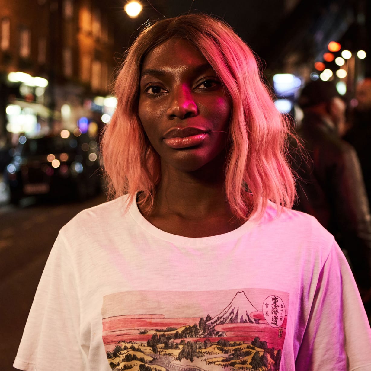 A woman with pink hair standing on a city street at night.