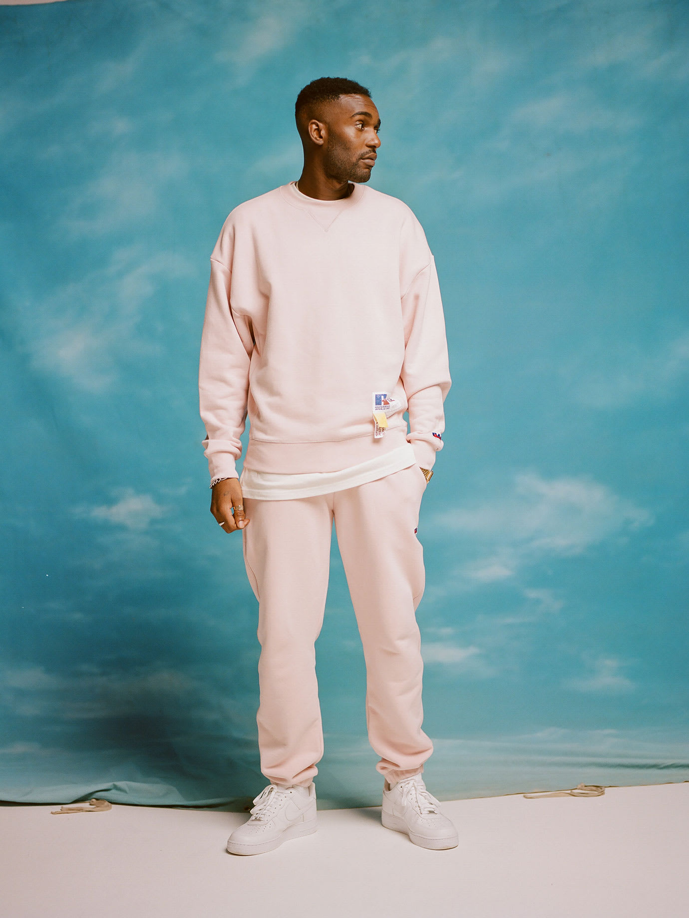 A man wearing pink sitting in front of a blue sky and clouds backdrop.