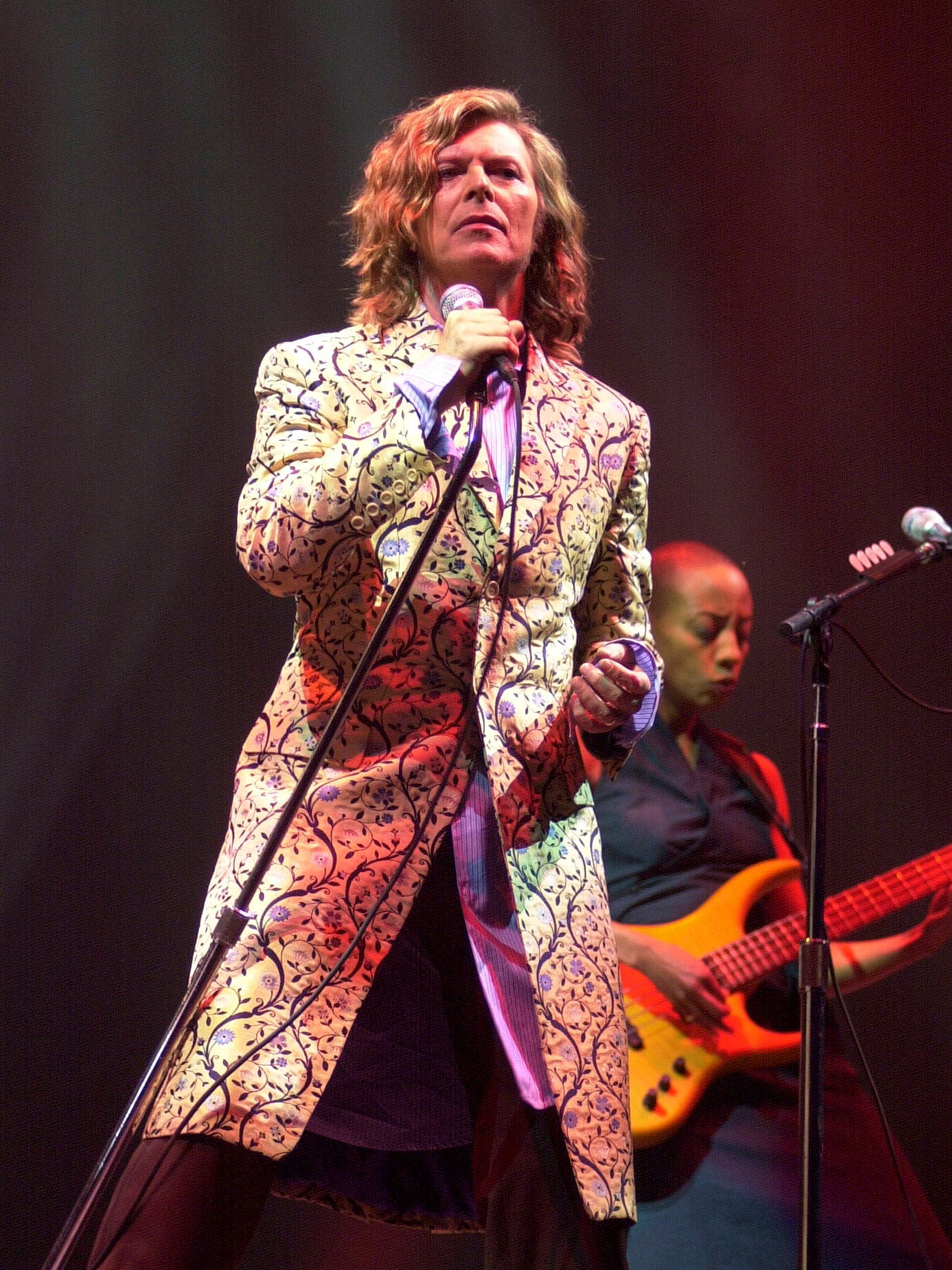 A man singing on a stage in a long coat with long hair.