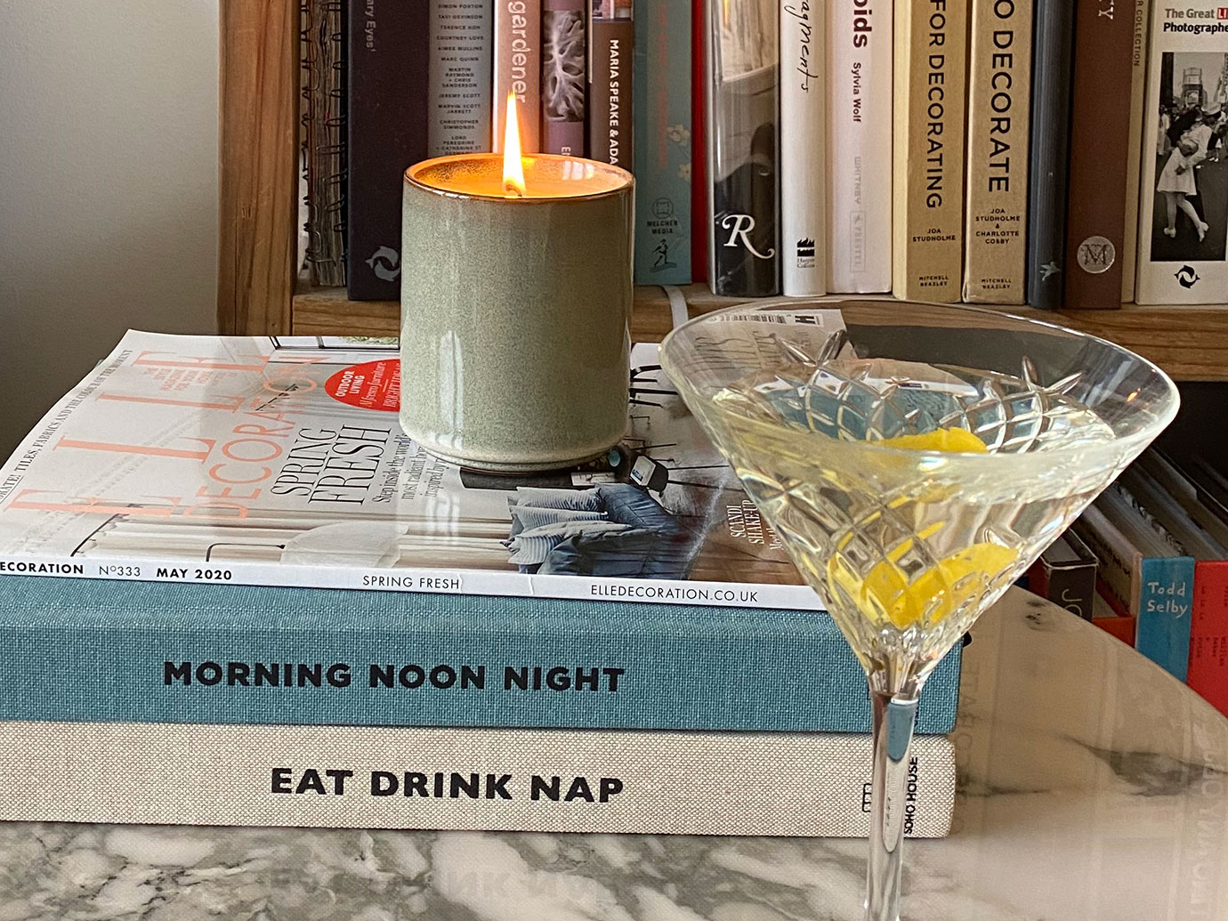 Two books on a table next to a cocktail and a lit candle.