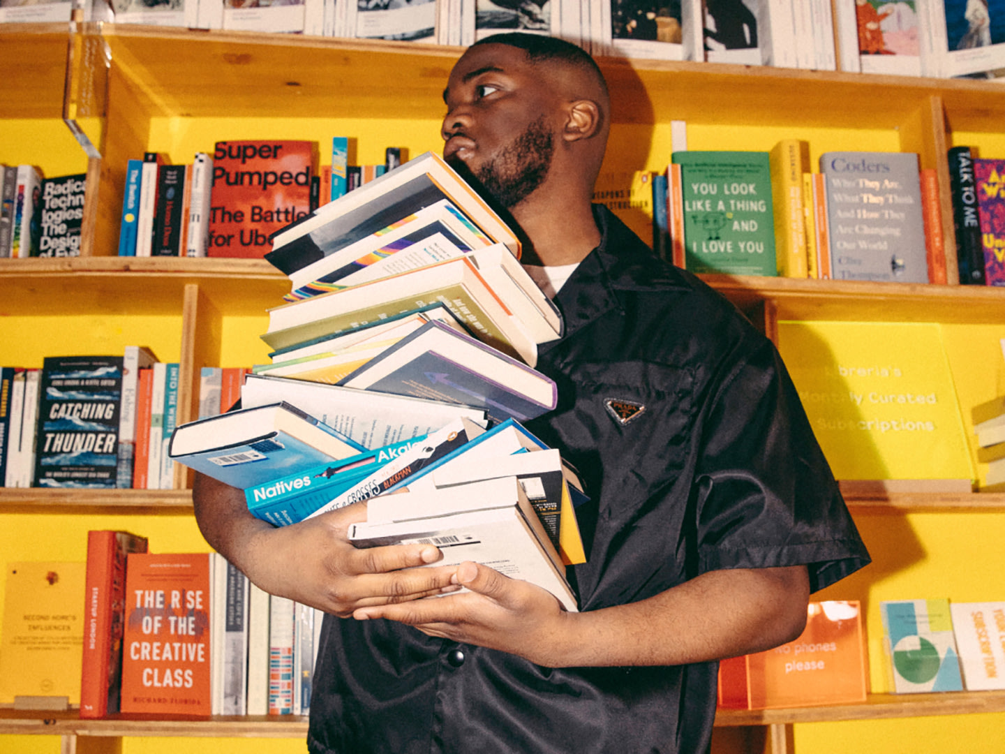 A man holding a pile of books next to a yellow bookshelf.