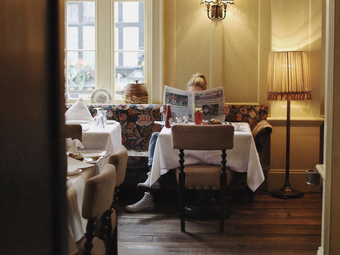 A woman reading a newspaper at a table in a yellow dining room.