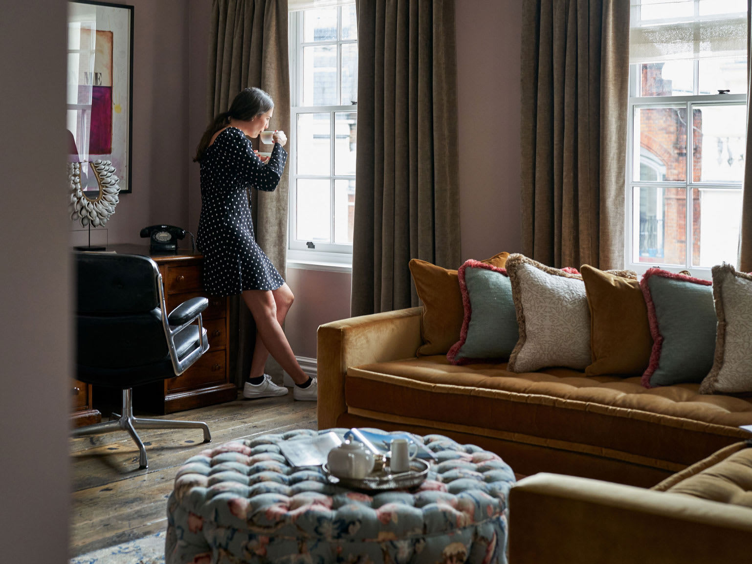A woman drinks a cup of tea in a lounge area while looking out a window.