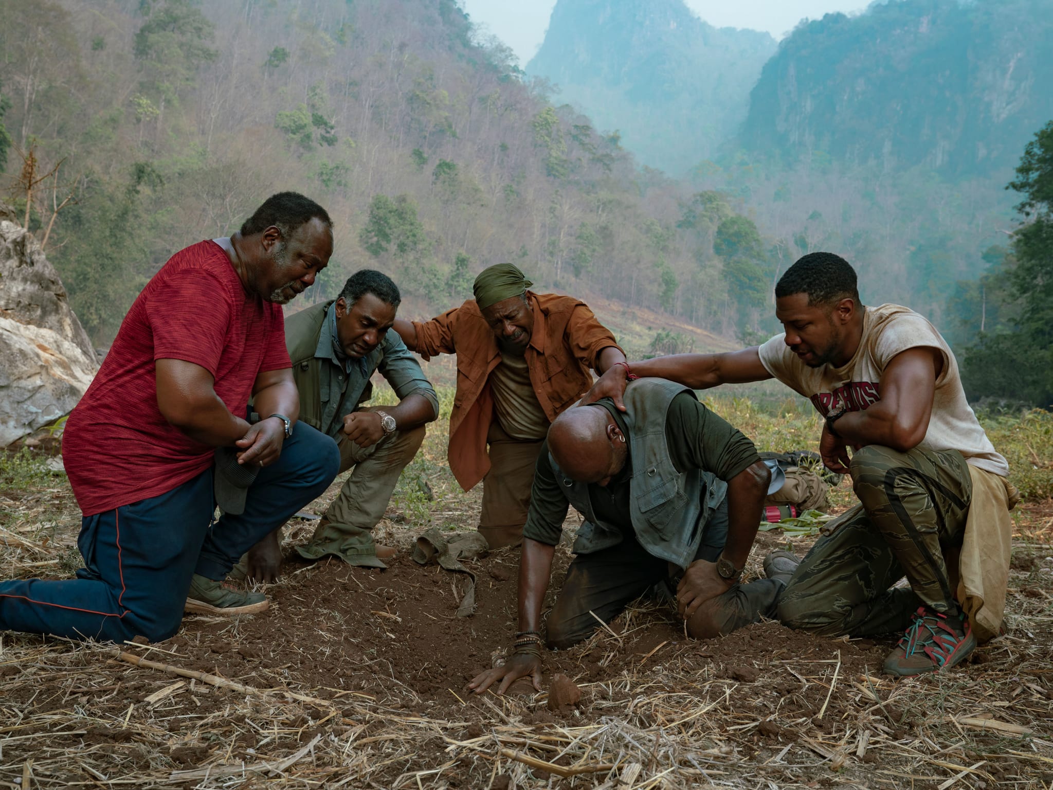 Five men with their arms around each other kneeling by a patch of ground.