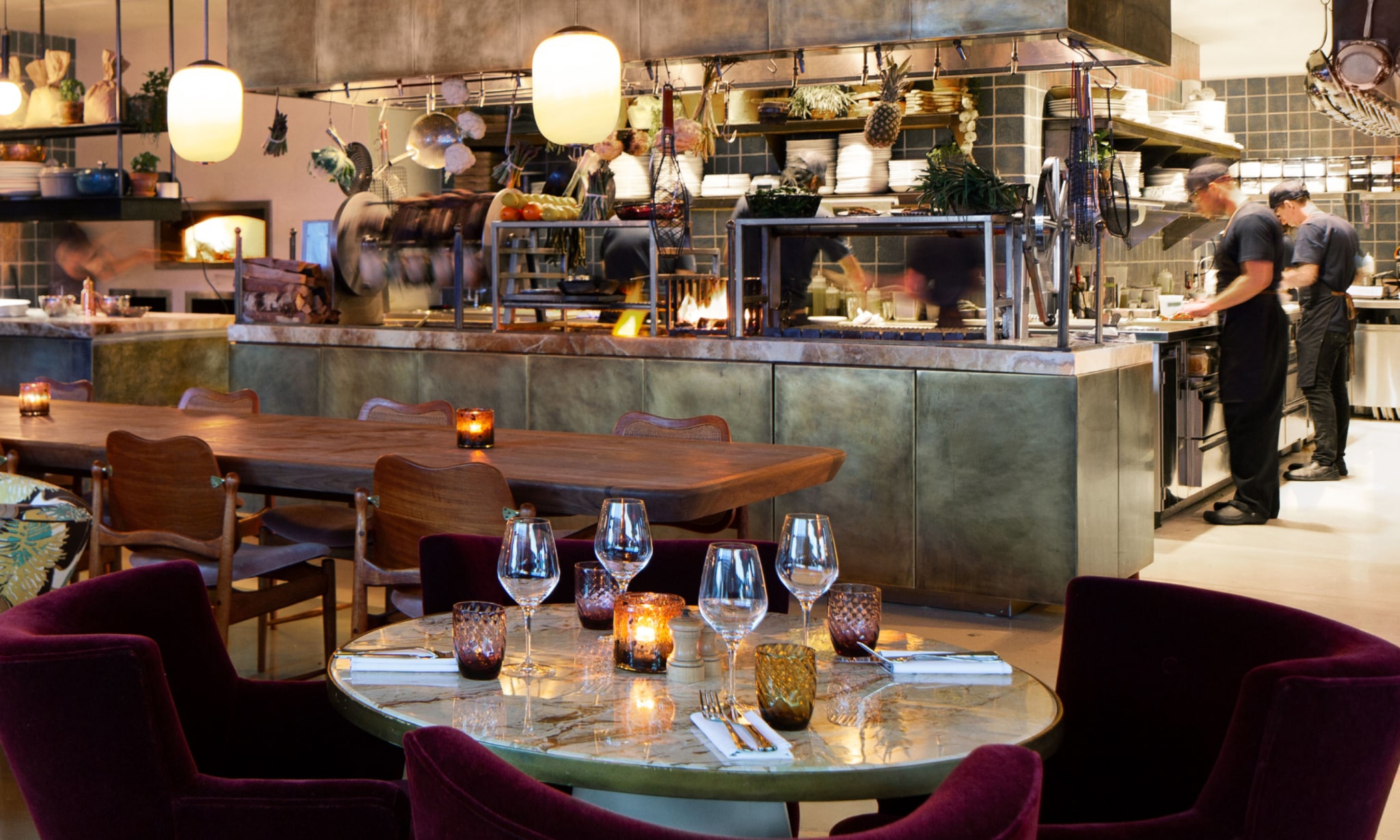 Chefs prepare food in an open plan kitchen next to a restaurant furnished in a relaxed style.