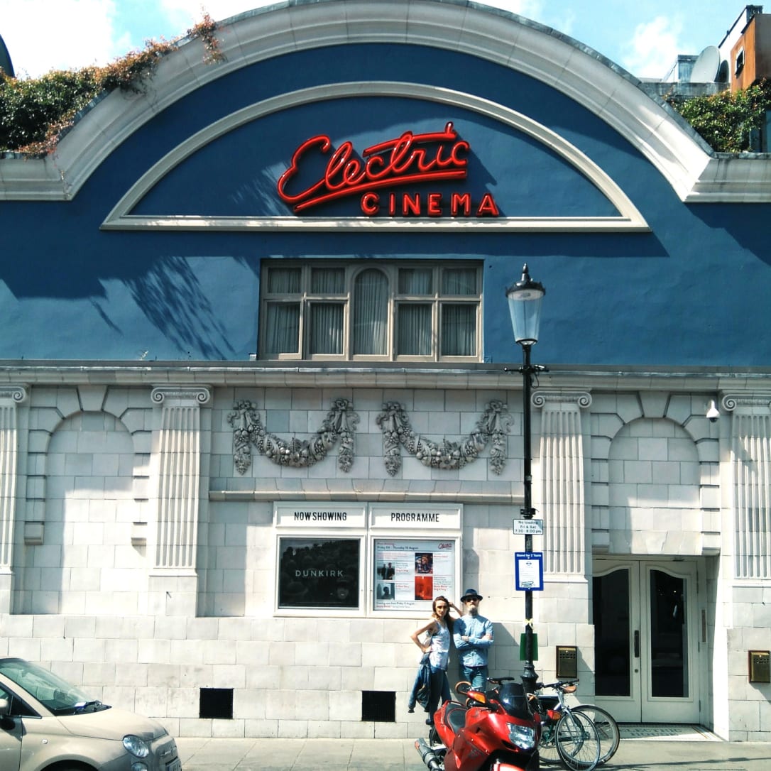 Two people standing outside the ornate facade of an old cinema with Electric Cinema written in neon at the top.