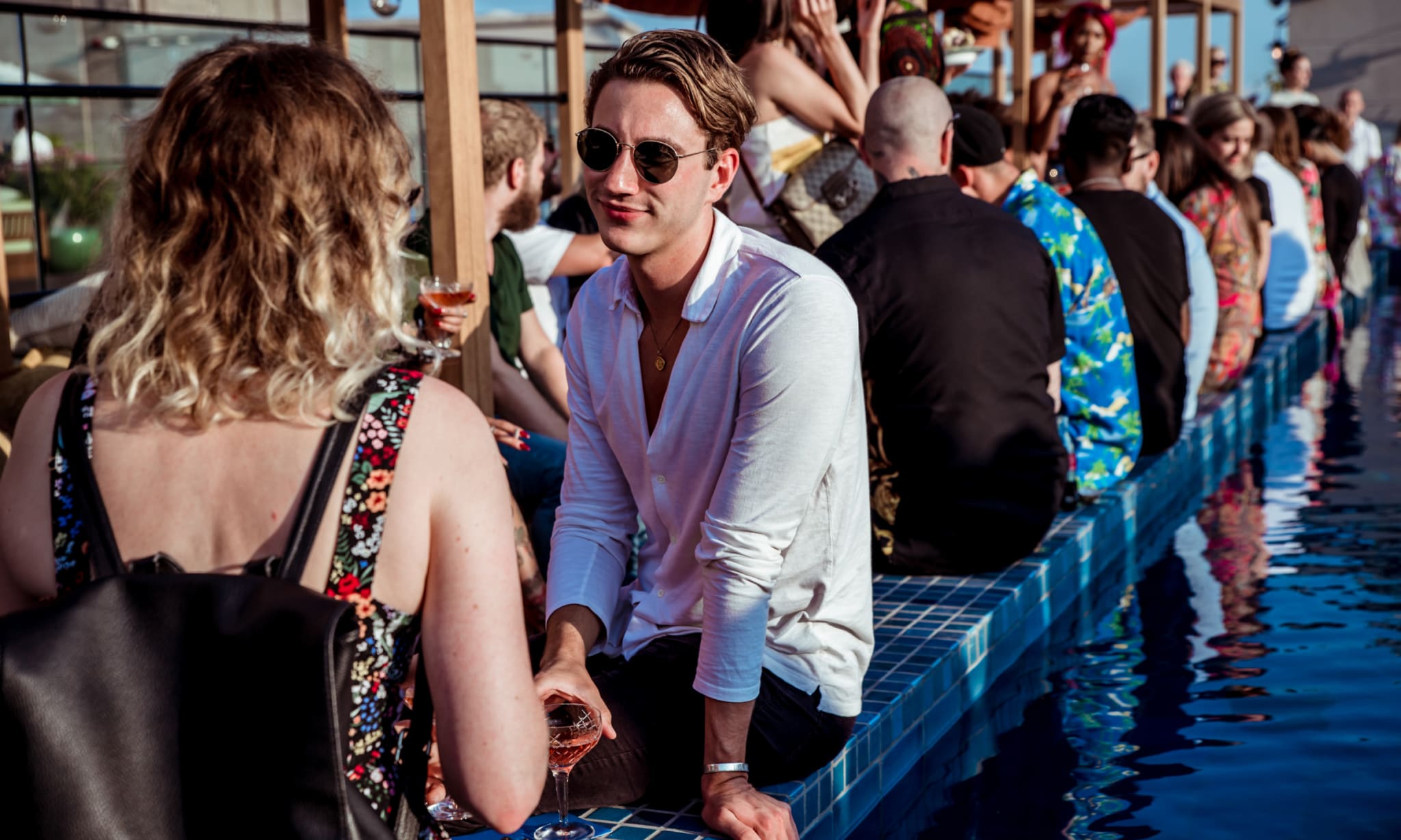 A group of people sat around a rooftop swimming pool on a sunny day.