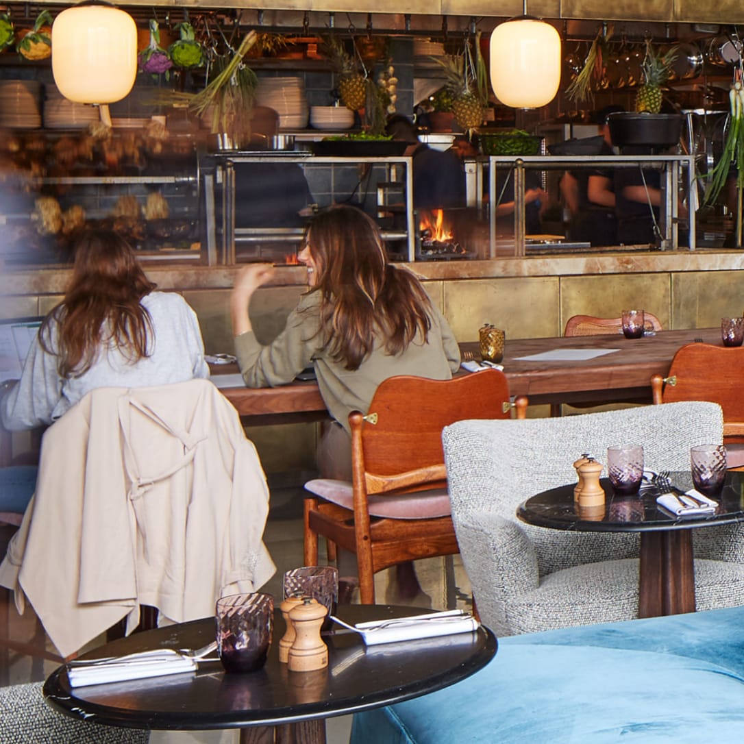 Two female diners chat while sitting at a table in a restaurant with an open plan kitchen.