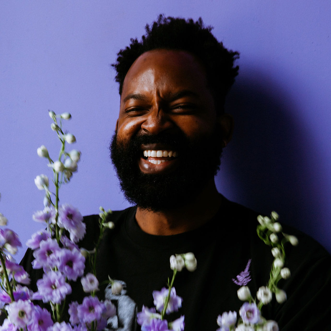 A man smiling in front of a purple background while holding flowers.