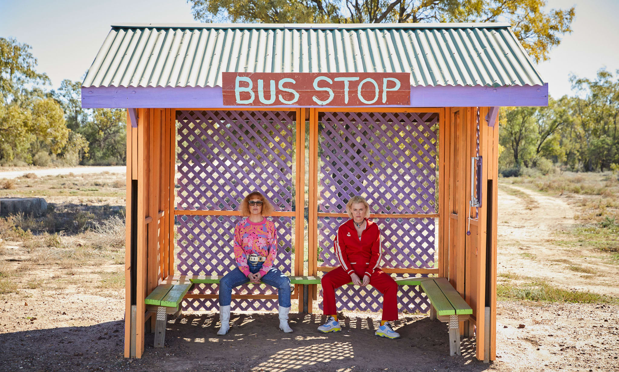Two women sat in a colourful bus stop in the desert.