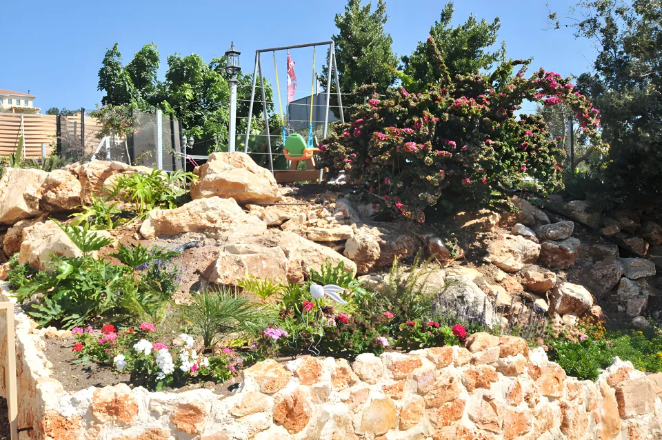 גינת סלעים פורחת בחצר של וילה לוקה, ומעליה נדנדות ילדים