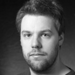 A black-and-white close-up portrait of a man with short, slightly messy hair and a beard. He has a calm expression and is looking directly at the camera, with a softly blurred dark background.