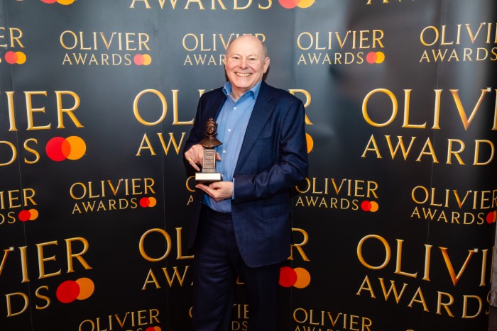 Robert Israel holds his Olivier Award statuette in front of a branded backdrop
