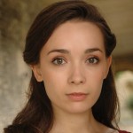 A close-up portrait of a young woman with long dark hair, soft makeup, and expressive brown eyes. She has a calm, neutral expression, with a blurred outdoor background.