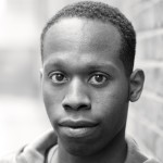 A black-and-white close-up portrait of a man with short hair. He has a neutral expression and is looking directly at the camera, with a softly blurred urban background.