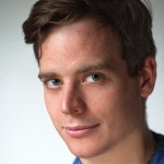 A close-up portrait of a man with short brown hair and blue eyes. He has a slight smile and is wearing a casual blue shirt, with a plain light background.