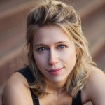 A close-up portrait of a young woman with wavy blonde hair and bright blue eyes. She has a gentle smile and is wearing a sleeveless black top, with a blurred outdoor background.