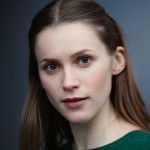 A close-up portrait of a young woman with long, straight brown hair and green eyes. She has a neutral expression and is wearing a green top, with a soft, dark background.