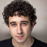 A close-up portrait of a young man with dark curly hair and blue eyes. He has a neutral expression and is wearing a black top, with a softly blurred background.