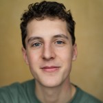 A close-up portrait of a young man with short curly hair and blue eyes. He is smiling softly and wearing a green top, with a neutral, softly blurred background.