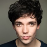 A close-up portrait of an actor with tousled dark hair and hazel eyes. They have a neutral expression and is wearing a dark top, with a softly blurred background.