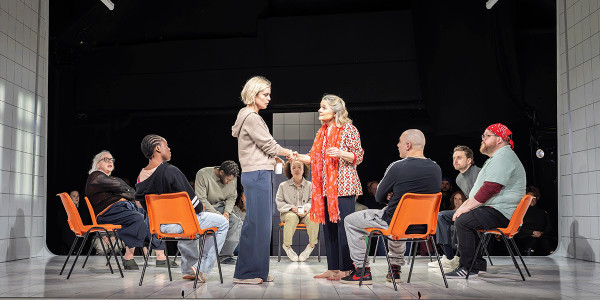 The cast are sat on bright orange chairs in a circle. In the centre Emma and the Therapist are shaking hands.