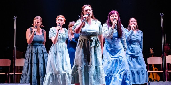 The cast of Spring Awakening 15th Anniversary Concert 2024 stand on stage wearing dresses in shades of blue, holding microphones.