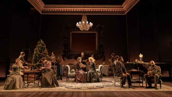 A group of actors sit on a stage in Victorian dress.