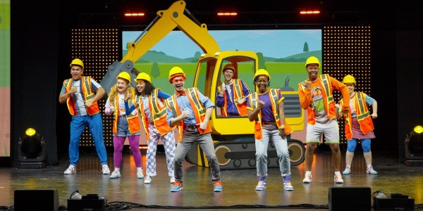 This image shows a lively stage performance featuring a group of people. They are all wearing construction-themed outfits, including hard hats and high-visibility vests, and are dancing in front of a large, cartoon-style backdrop of a digger. The stage is well-lit, and the background includes animated scenery. The central figure, dressed in an orange bow tie and hat, is Blippi.