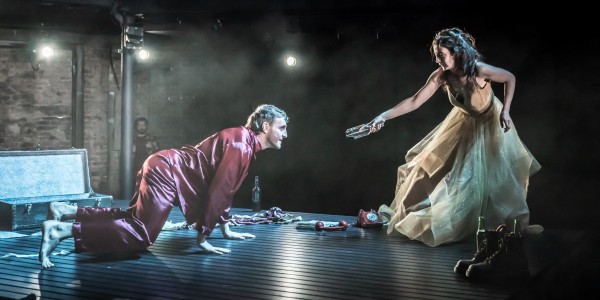 Paul Mescal, kneeling in red pyjamas, looks up at Patsy Ferran, dressed in a gold gown, as she leans forward, handing him a bottle in a dramatic stage scene. The dim lighting and sparse set add to the tense atmosphere.