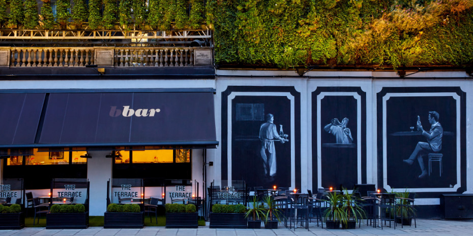 Outdoor seating area of a bar named "bbar" with a terrace sign, green wall above, and three black-and-white murals depicting various scenes on the facade.