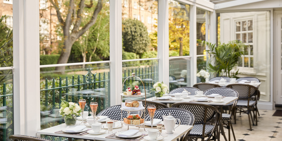 Sunlit conservatory with white tables, wicker chairs, and a view of a garden. Tables are set with teacups, saucers, and a three-tiered stand of pastries and sandwiches.