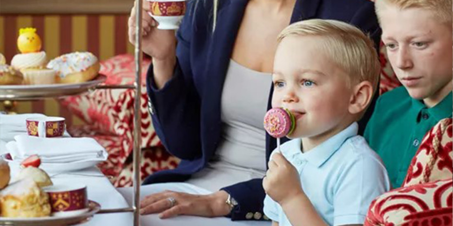 A young child with blonde hair, dressed in a white polo shirt, enjoys a cake pop while sitting next to an older blond boy in a green shirt. Across the table, pastries and sweets are displayed on a tiered stand. An adult seated with them holds a teacup.