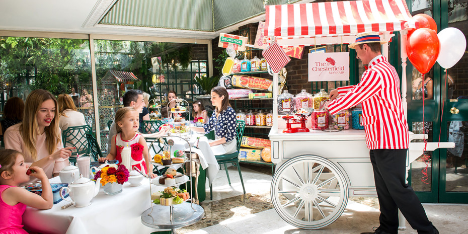  A man in a red and white striped jacket serves sweets from a cart at The Chesterfield Mayfair. Families with children are seated at tables, enjoying tea and snacks. The room is well-lit with natural light and decorated with colorful balloons.