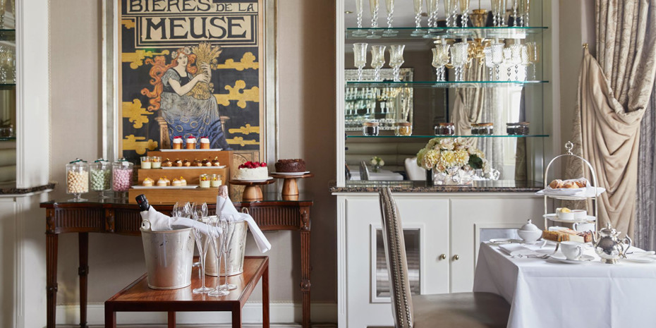 A cozy dining room with a vintage "Bieres de la Meuse" poster on the wall. A table features desserts, including cupcakes and a cake, with a bucket of champagne nearby. Another table is set with a white tablecloth, tableware, pastries, and a tea set. Shelves display glassware.