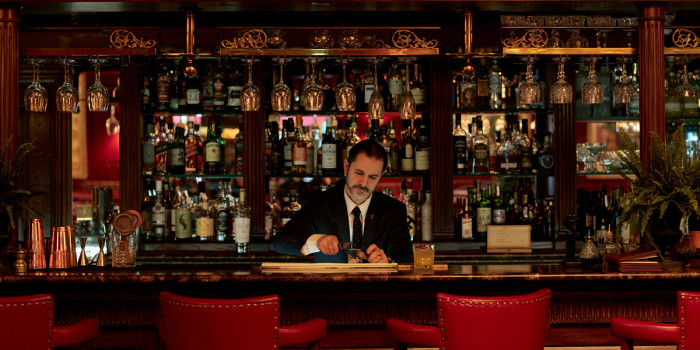 A staff member prepares a drink behind a bar with red seats in front and a large selection of drinks in glass bottles visible behind