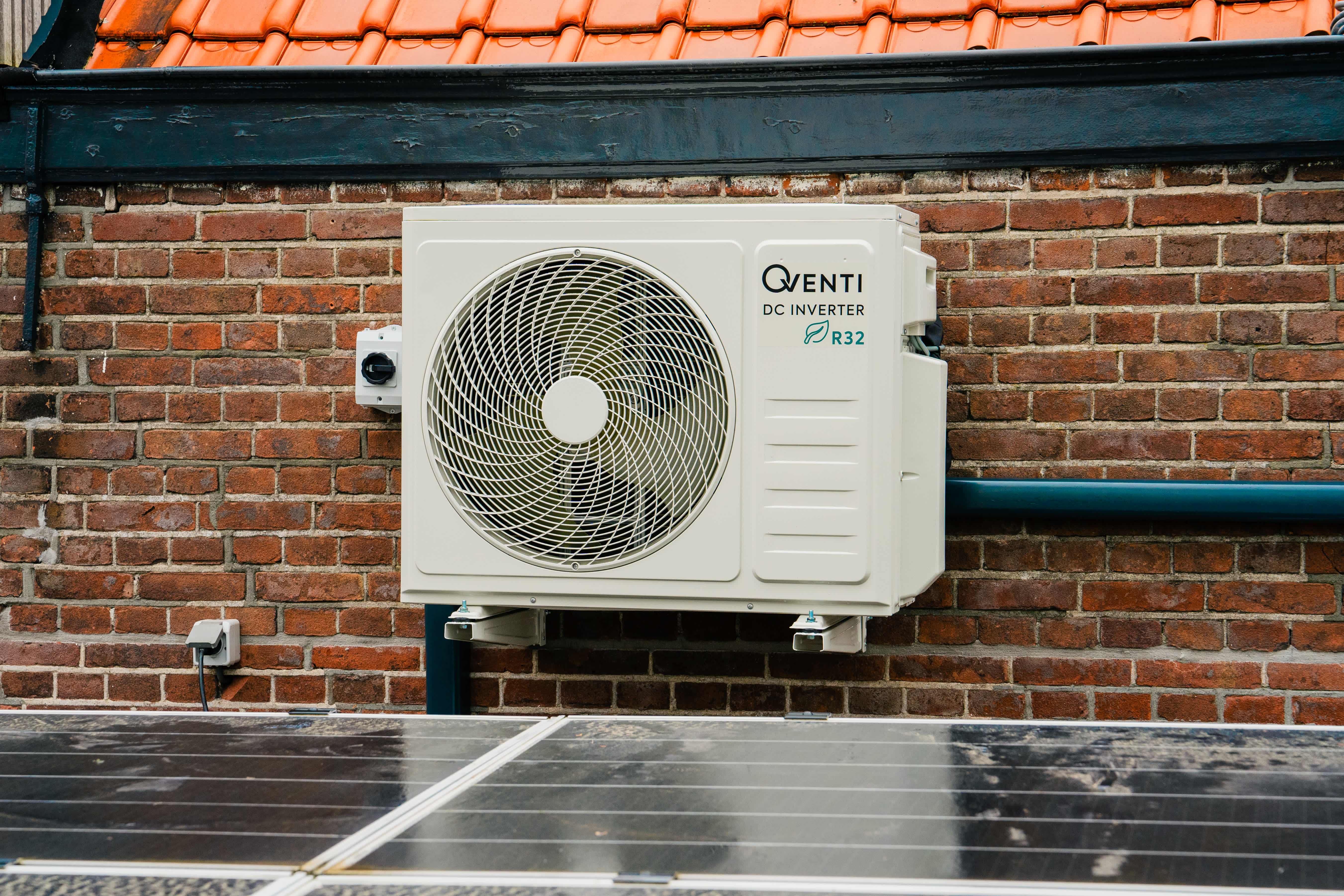 Udendørsenhed til en varmepumpe monteret på en murstensvæg, med solpaneler i forgrunden