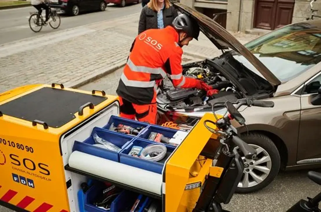 Autoredder står ved bil med motorhjelm åben. Ved siden af bilen holder vejhjælpscykel. 