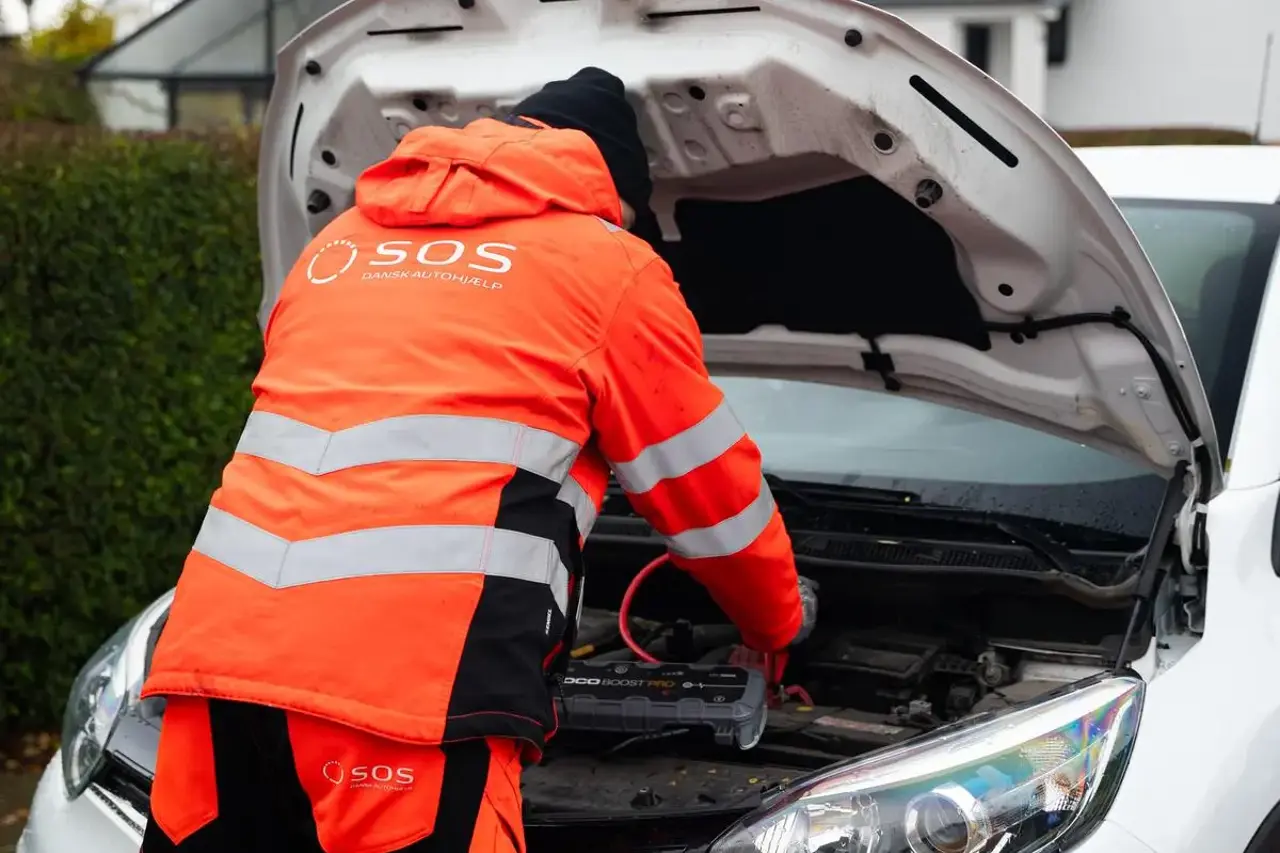 Autoredder står med rykken til og er i gang med at starte bil. Motorhjelm er åben og der er påsat booster til batteriet på bilen. 