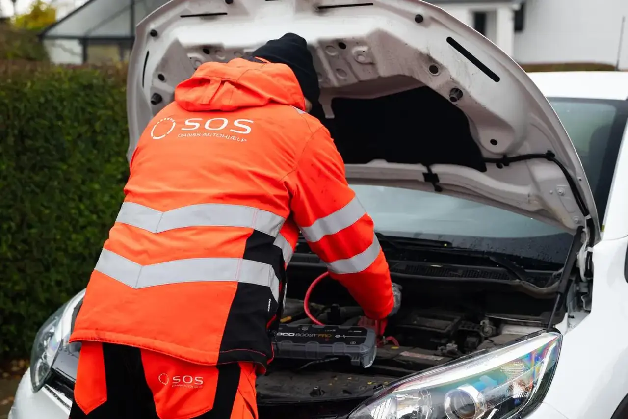 Autoredder står med rykken til og er i gang med at starte bil. Motorhjelm er åben og der er påsat booster til batteriet på bilen. 