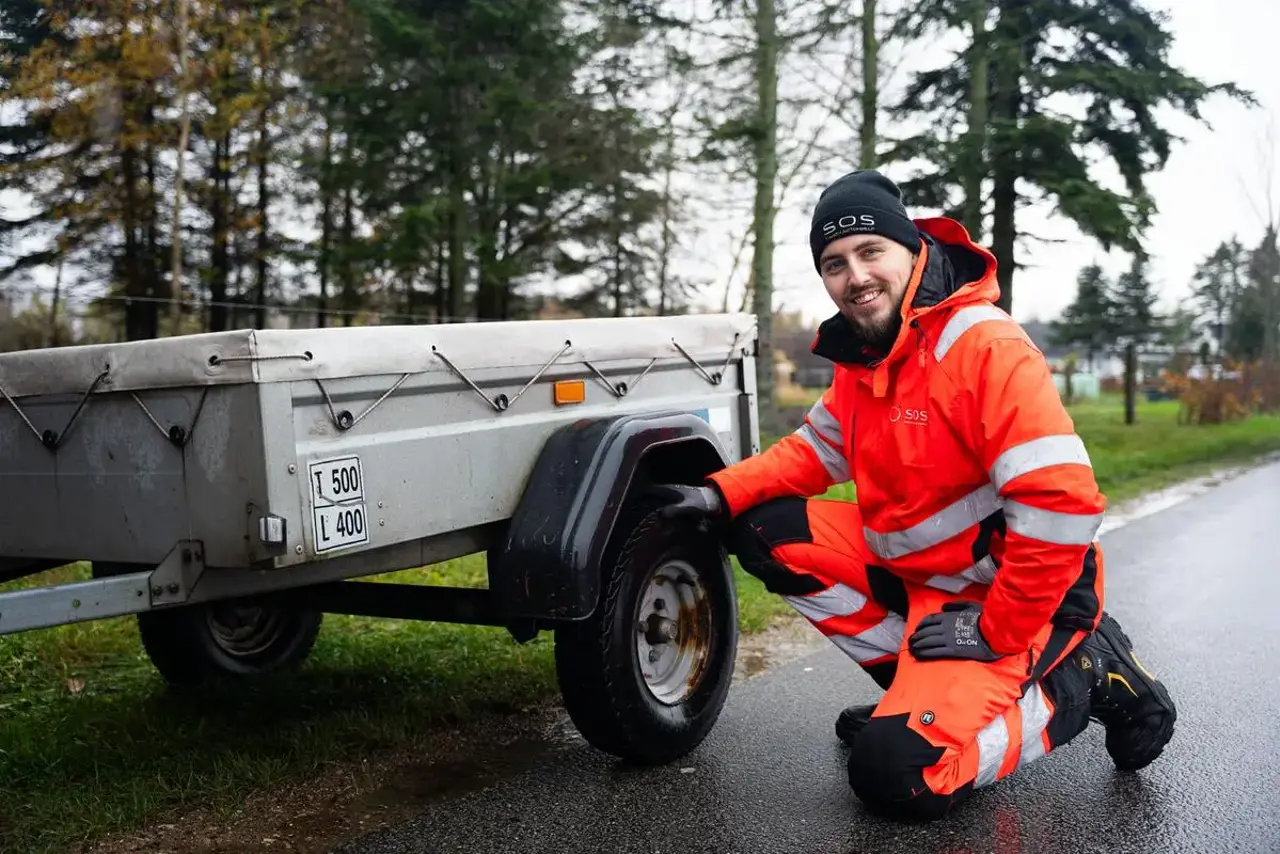 Autoredder sidder på huk ved trailer og smiler
