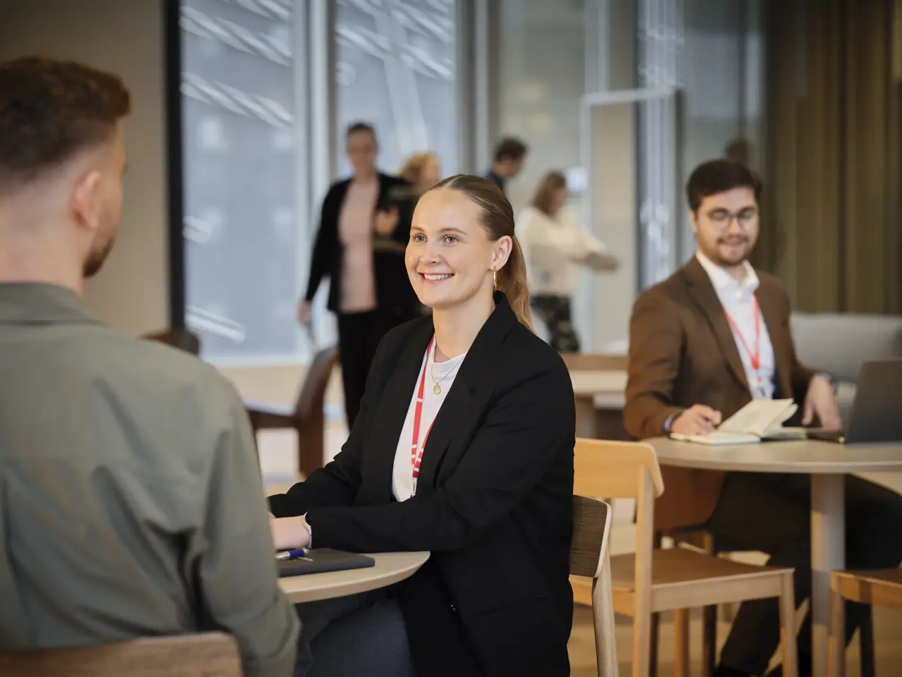 Pige der smiler ved et bord. Mand sidder med ryggen til kameraet. Mand i baggrunden.