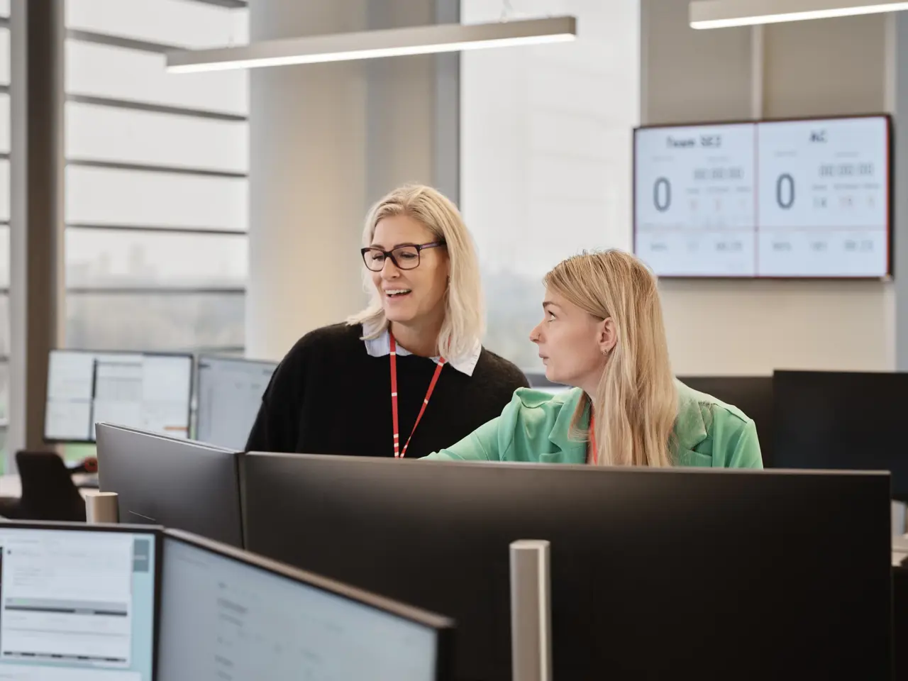 2 kvinder foran computer skærme i en alarmcentral. Skærm i baggrunden på væg. 