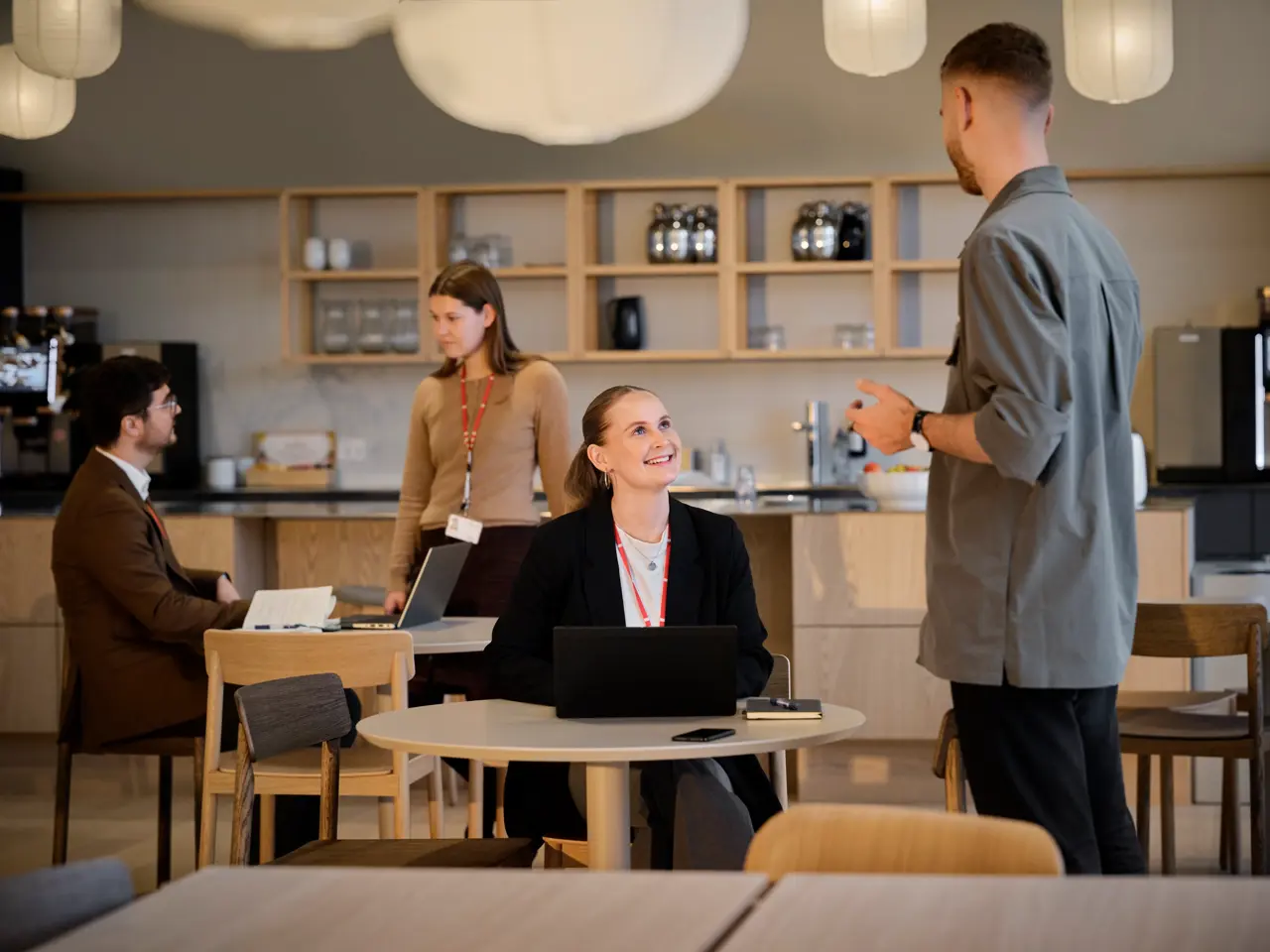Fyra personer i kök på arbetsplatsen. Flicka sitter vid bordet och tittar upp på en man som står.